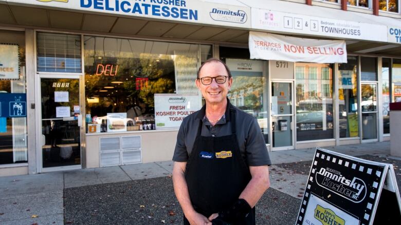 A man with glasses stands in front of a deli.