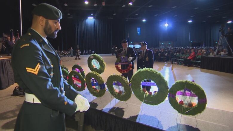 A man in a military uniform stands in front of a row of several wreaths, with a large crowd of seated people in the background.