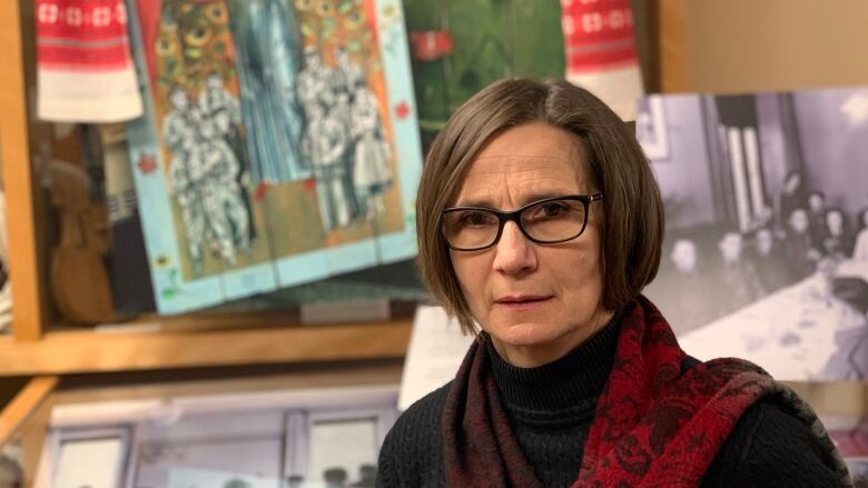 A woman with hair cut in a bob wears a black turtleneck and a scarf. She stands in front of a museum exhibit of Ukrainian folk art.