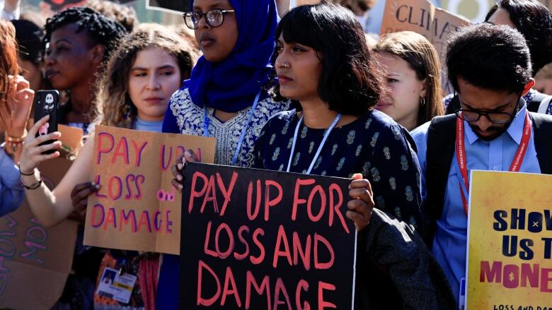 A young woman with shoulder-length brown hair and a nose ring stands at the front of a crowd of protesters, holding a sign that reads: 