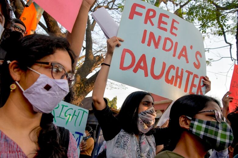 Three young women in face masks stand among a crowd of protesters. The one in the centre holds a sign above her head that reads: 