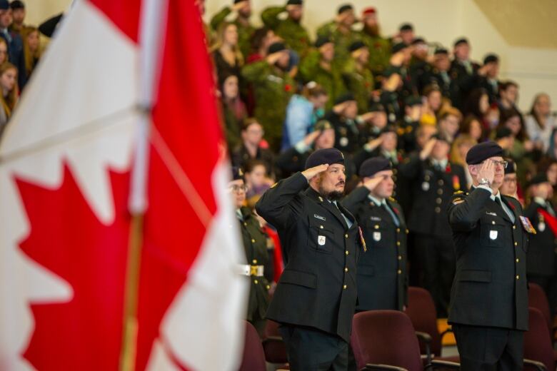 Soldiers in uniform stand and salute at a ceremony with a Canadian flag in the foreground.