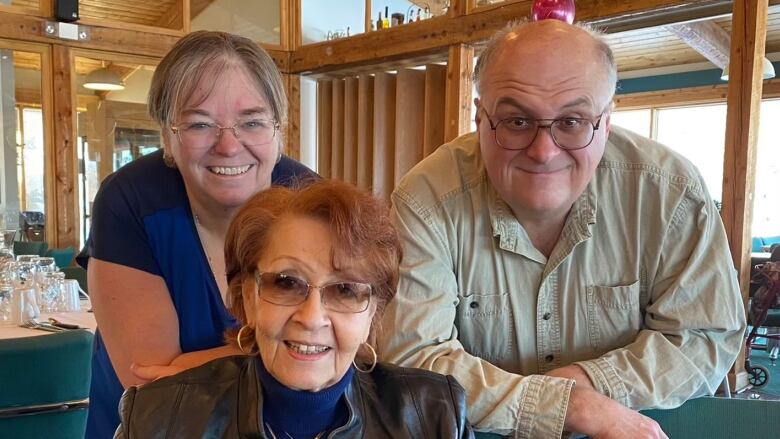 A man and a woman lean on the back of a chair an older woman is sitting in and smile.