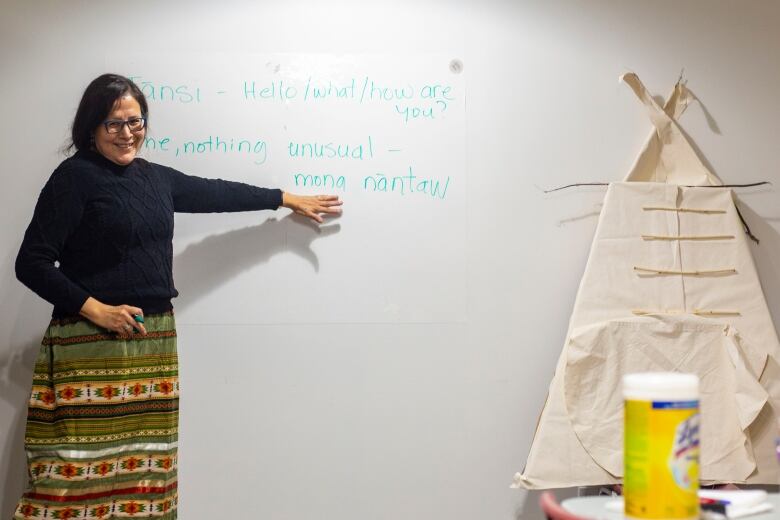 A woman wearing an ribbon skirt smiles while teaching in front of a white board with Cree words.