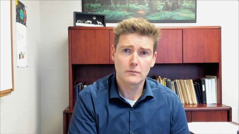 A man with brushed up light brown hair sits in an office. There's a brown cabinet with standing folders behind him.