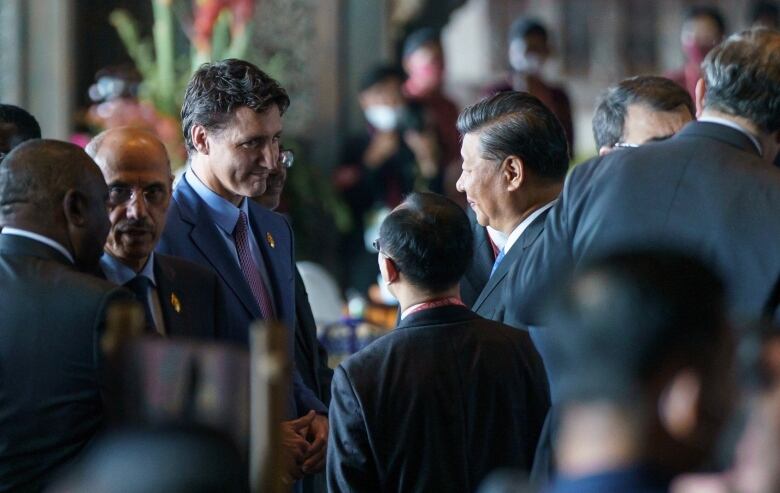 The Canadian prime minister smiles at the Chinese leader, who is accompanied by an aide. In the background, several individuals speak to each other.