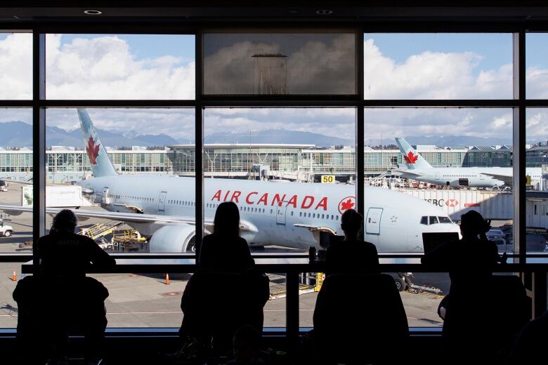 A row of people, in silhouette, wait at an airport departure lounge with an Air Canada plane visible in the background.