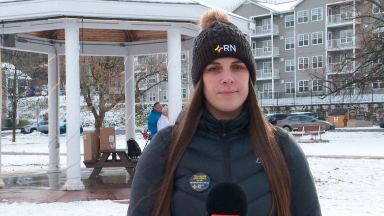 A woman wearing black jacket and black toque stands in front of a gazebo. The ground is covered in a dusting of snow.