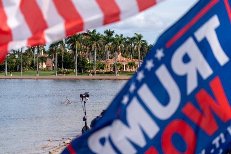 A large home and property is seen from across a body of water, with a Trump flag in the foreground. 