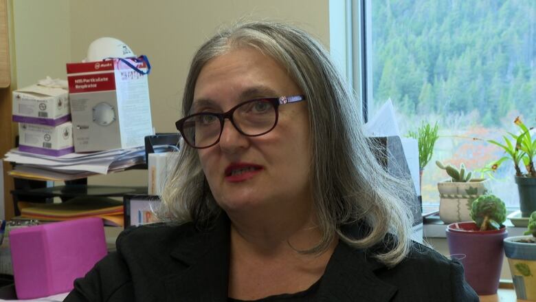 A woman with grey hair down to her shoulders sits at a desk in her university office. She's wearing a black shirt with dark red glasses.