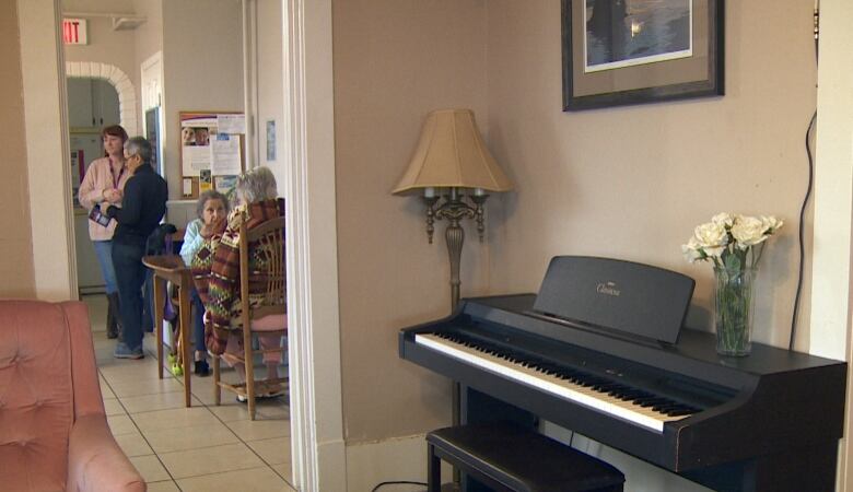 An electric piano placed against a pinkish-beige wall can be seen in the foreground, next to a doorway opening into a dining room, where two people are eating at a table, and another two are chatting as they lean against a wall.