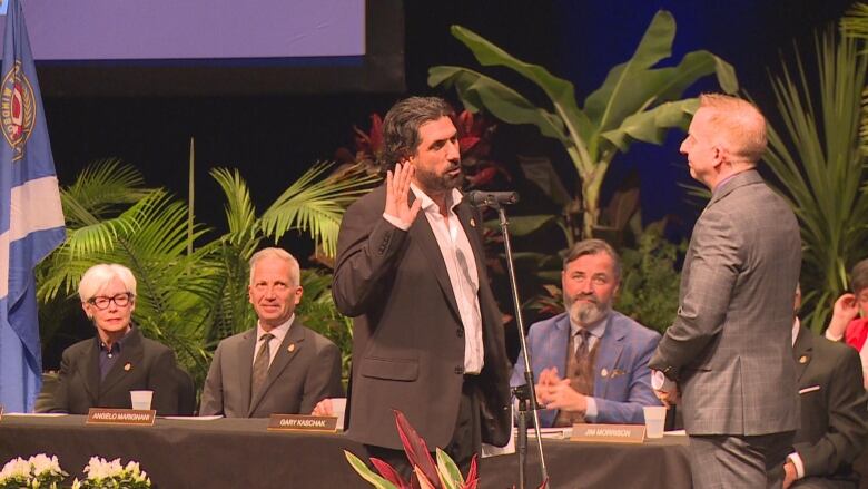 A man with medium-length hair and a beard raises his right hand on a stage. Sitting behind are other councillors. 