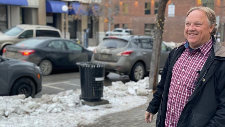 A middle-aged man in a red and white plaid shirt and black winter coat stands smiling on a sidewalk, with a small bank of dirty snow behind him.