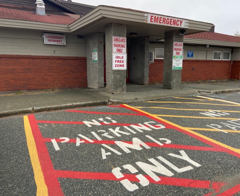 An sign that says emergency in red lettering hangs over the doors on a brick building with concrecte pillars. Signs  on the pillars say ambulance parking only.