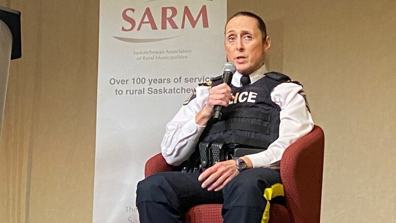 A woman in a police uniform speaks into a microphone as she sits in a chair. Behind her is a banner reading 