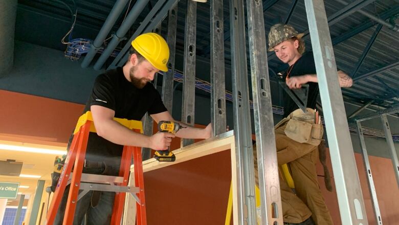 Two men work dressed in construction work gear work on a metal and wood wall structure.