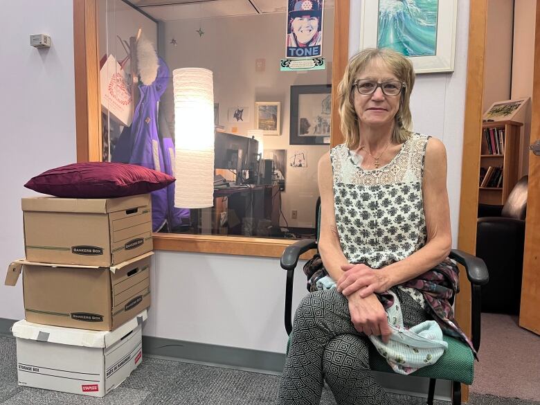 A woman sits on a chair in a lobby outside an office. Through the window behind her, you can see a desk, books and lamp. She's a middle-aged woman with blonde hair and glasses with a neutral expression.