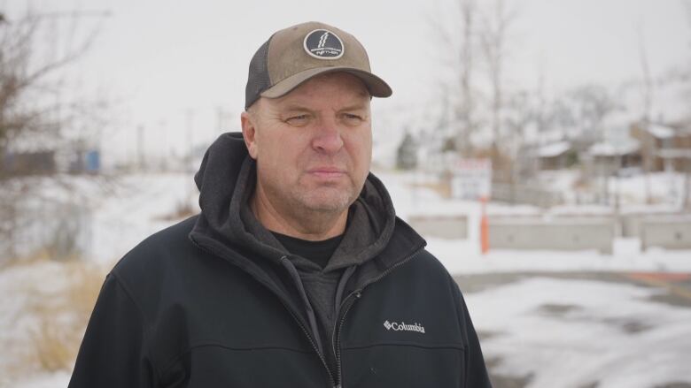 A white middle aged man wears a black jacket, a tan baseball hat and a scowl. He is photographed from the shoulders up standing outside with snow in the background.