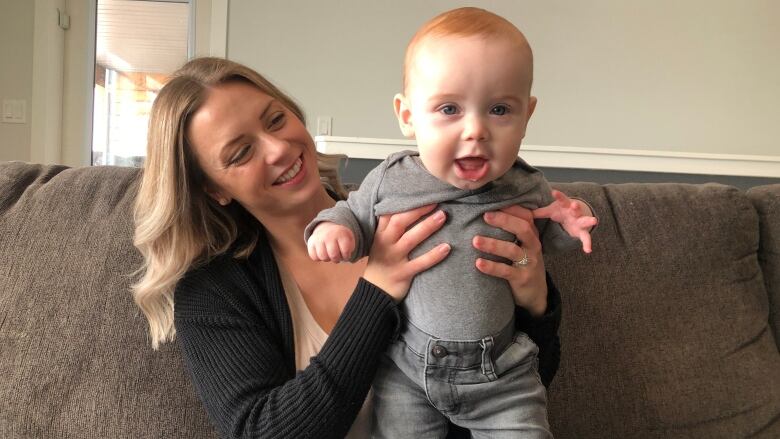 A smiling woman sits on a couch, holding up a smiling baby.