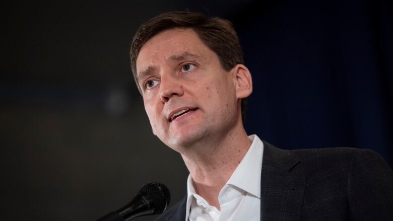 David Eby is photographed from the chest up. He stands in front of a black background at a microphone wearing a black jacket and white shirt with no tie. He has sandy-coloured brown hair and is clean shaven. His mouth is slightly open as if about to speak.