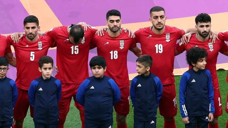 Men wearing red jerseys stand shoulder to shoulder, arms links, with solemn faces, and children wearing blue jerseys stand in front of them. 