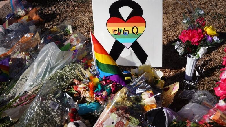 A pile of flowers, stuffed animals and rainbow flags on the ground surrounding a small handmade sign with the Cub Q logo: A rainbow heart over a black ribbon, with the words: 