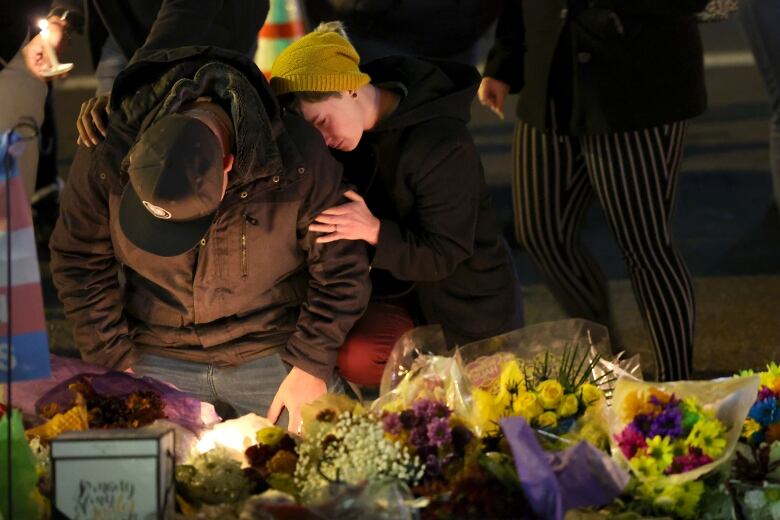 Two people, clad in winter gear, kneel outside at night in front of a makeshift memorial with flowers and candles. One person has their arms wrapped around the other in an embrace. 