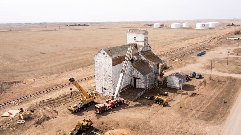 An old wooden elevator, painted white, is seen on baren prairie land surrounded by trucks and other equipment.