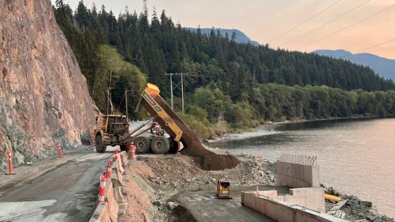 A dump truck pours a load of dirt off the side of a dirt road down an embankment. There are evergreens and a lake in the background.