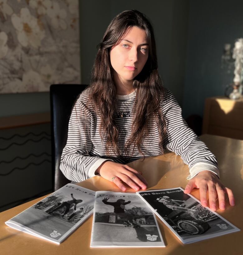 A woman with long dark hair in a black and white striped shirt sits at a table behind three self-published zines, all bearing the title 