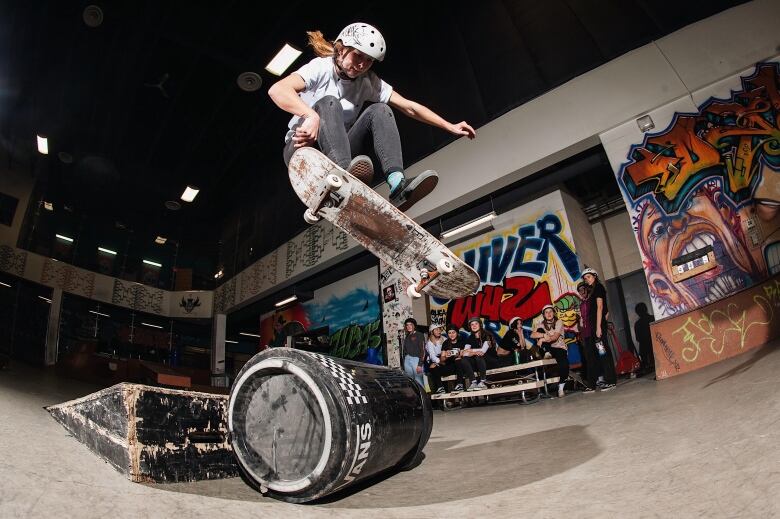 A person with long blonde hair wearing a white t-shirt does a skateboarding trick in an indoor skatepark, with a number of people watching in the background.