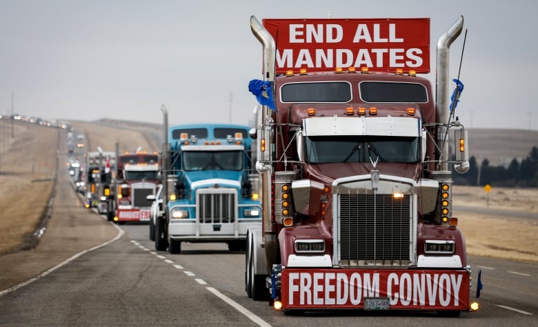 A line of semis can be seen on the highway, driving away from the protest. On top of the first truck sits a red and white sign reading 