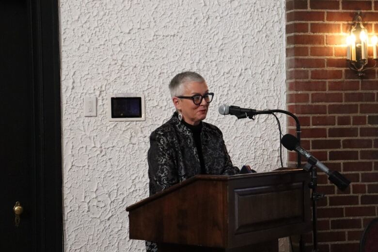 A woman with short grey hair and big black glasses speaks at a podium. 