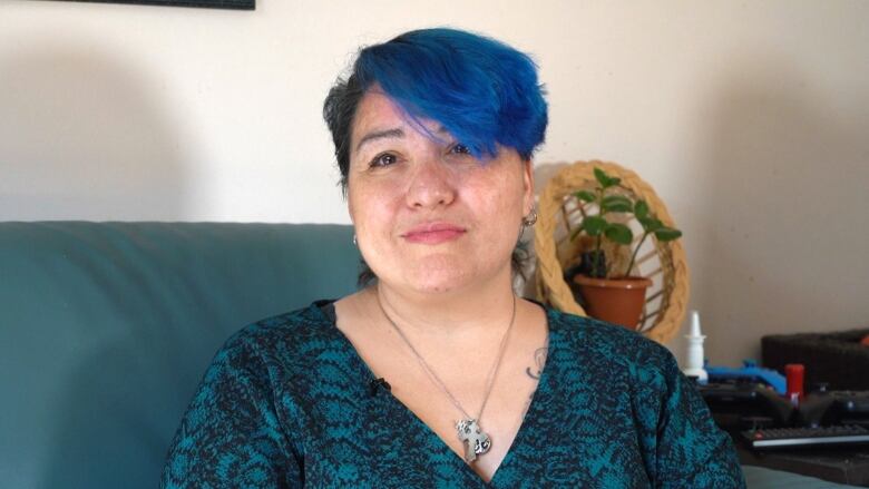 A woman with short dark hair dyed vivid blue on one side sits on a green sofa and faces the camera.