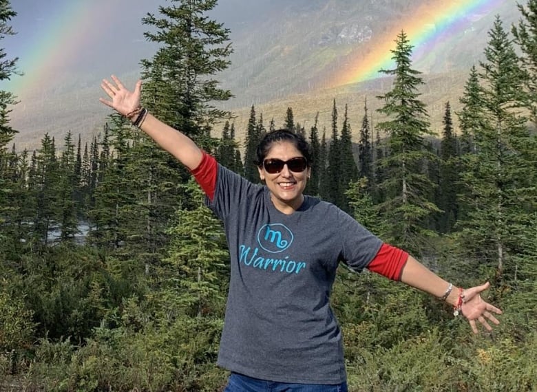 A woman with a huge grin stands on a rock in the mountains with a rainbow behind her.