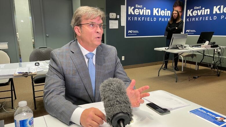 Kevin Klein sits at a table in front of a microphone, as a woman stands several feet behind him, in a corner where two of his Kirkfield Park campaign posters are plastered.