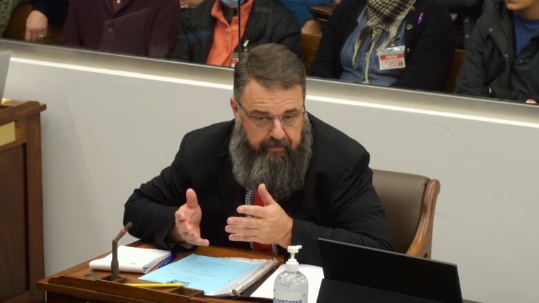 A man sitting at a desk, wearing a dark suit jacket and with a full beard, gestures as he speaks. 