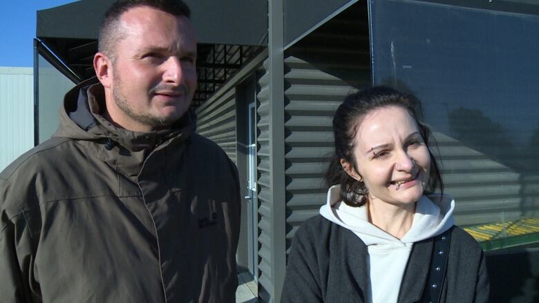 A man wearing a dark green jacket standing next to a woman with facial piercings wearing a white hoodie and a grey jacket