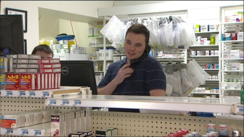 A man speaks on the phone. He is standing behind shelves at a store.