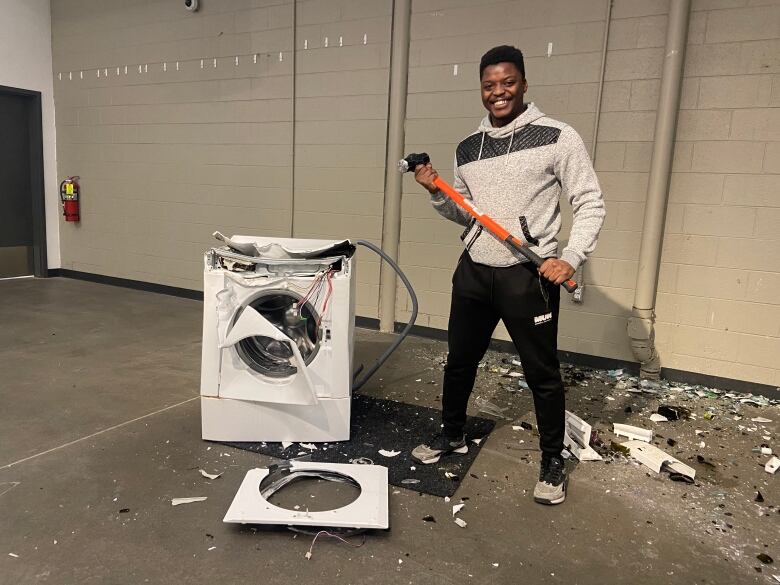 A man smiles while holding a sledgehammer after destroying a washing machine.