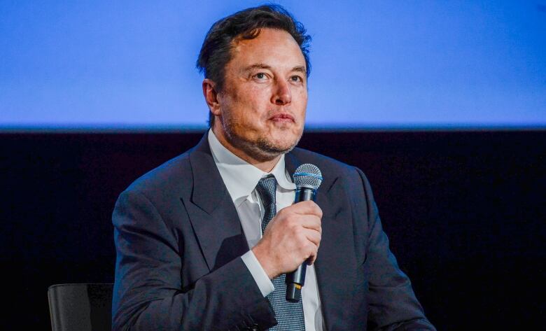 Elon Musk is wearing a suit and holding a microphone.