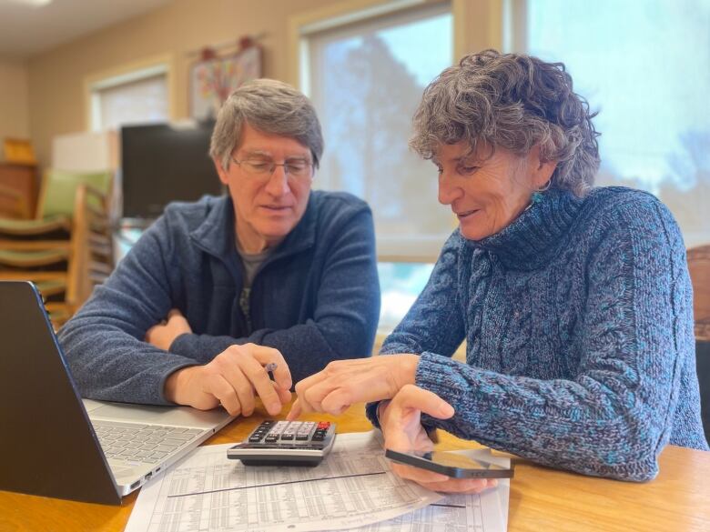 A man and a woman, both with grey hair, sit together at a table with papers and a laptop computer, punching numbers into a calculator. 