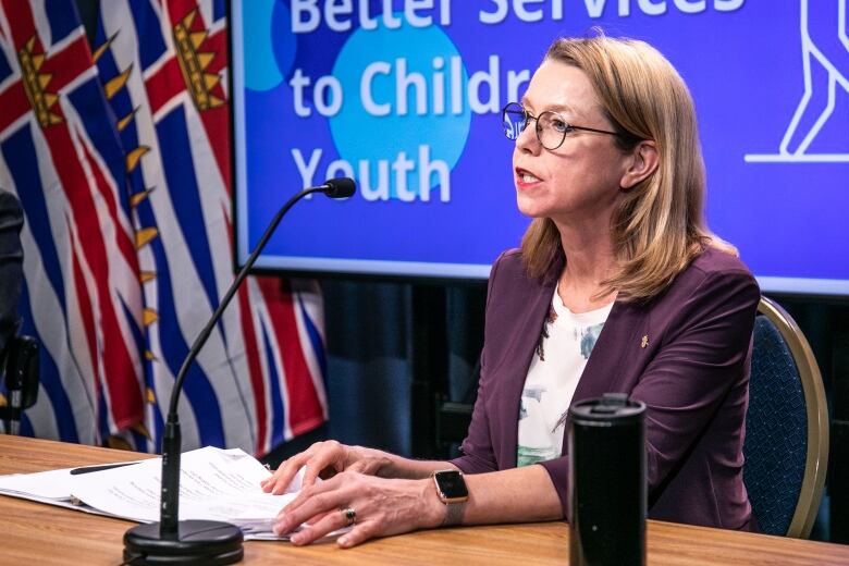 A white woman wearing a purple coat addresses a news conference, with a slide behind her reading 'Better services to children and youth.'