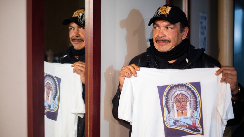 A man holds a t-shirt with an Indigenous art print while looking in a mirror.