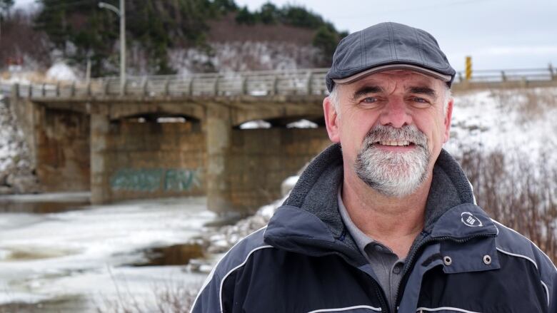 A bearded man stands in front of a bridge on a snowy day.