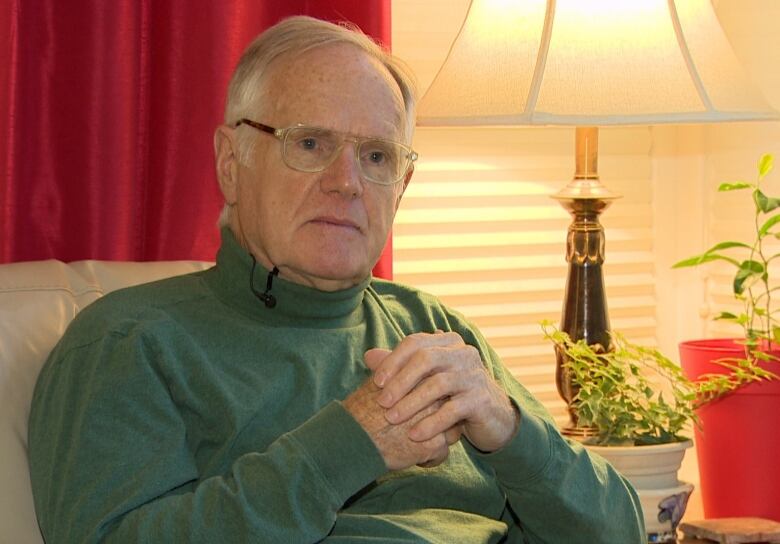 A person in a green turtle neck sits on a white sofa next to a lamp with his hands folded.