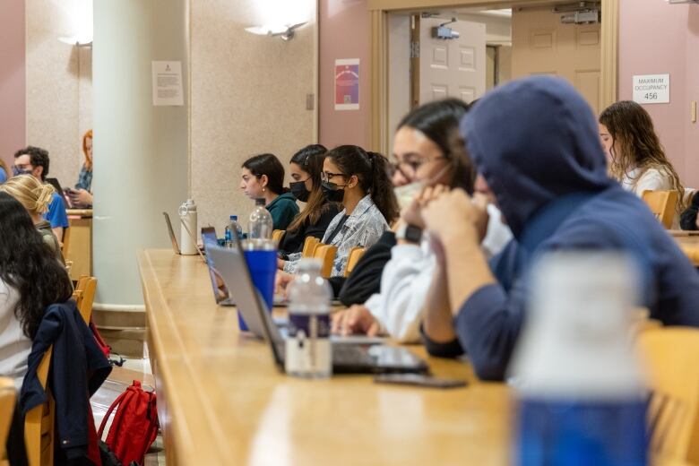 University students, some wearing masks and some not, are seen from the side typing notes on laptop computers as they sit at long wooden desks in a lecture hall.