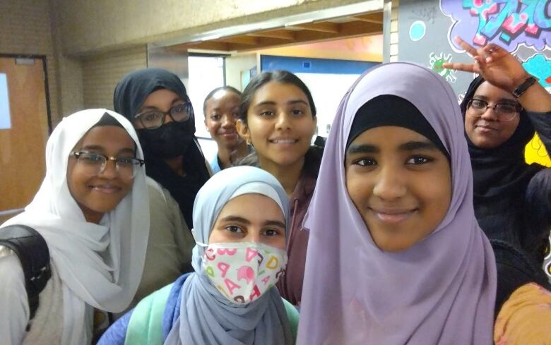 A group of seven smiling teen girls, more than half of whom are wearing hijabs, pose for a photo inside a school hallway.