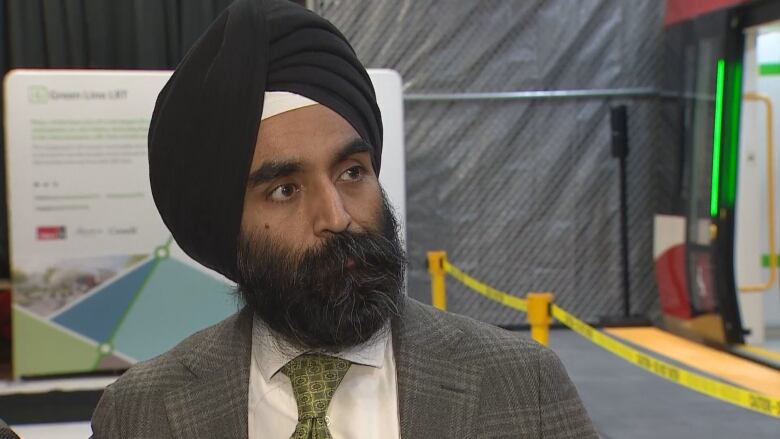 A man with a beard, a black turban, a gray suit and green tie stands before a red model of the light rail train. 