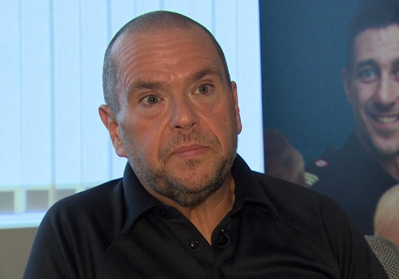 A shoulder-up shot of a person wearing a black polo shirt sitting in front of a window.